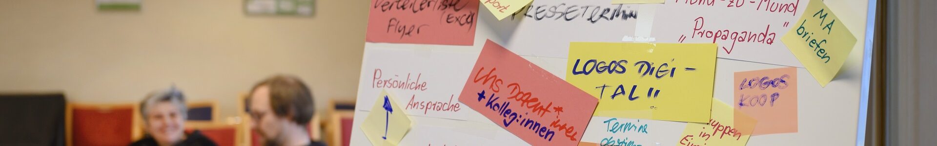 Im Vordergrund des Raumes steht eine Pinnwand mit Post-Its zu Ideen der Öffentlichkeitsarbeit. Im Hintergrund sitzen zwei Personen.