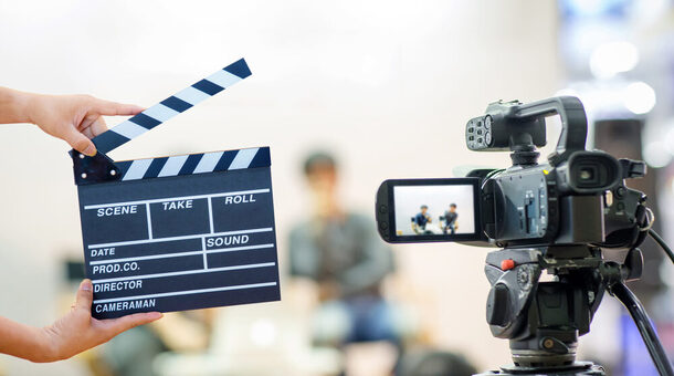Eine Videoaufnahmesituation wird dargestellt. Links ist eine Kameraklappe und rechts eine Videokamera. Der Hintergrund ist verschwommen.