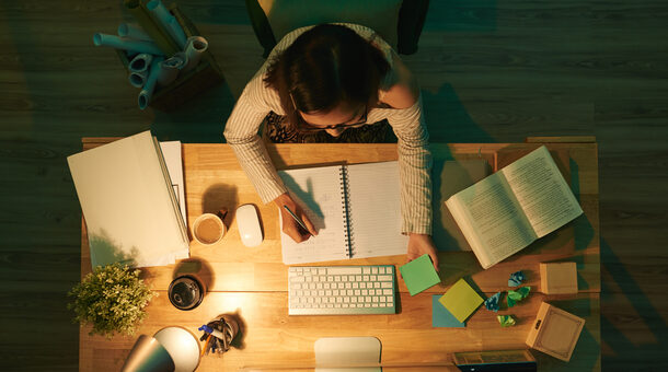 Bild von oben. Person sitzt am Schreibtisch, vor ihr liegen ein Block, ein Bildschirm, Bücher und Notizzettel.
