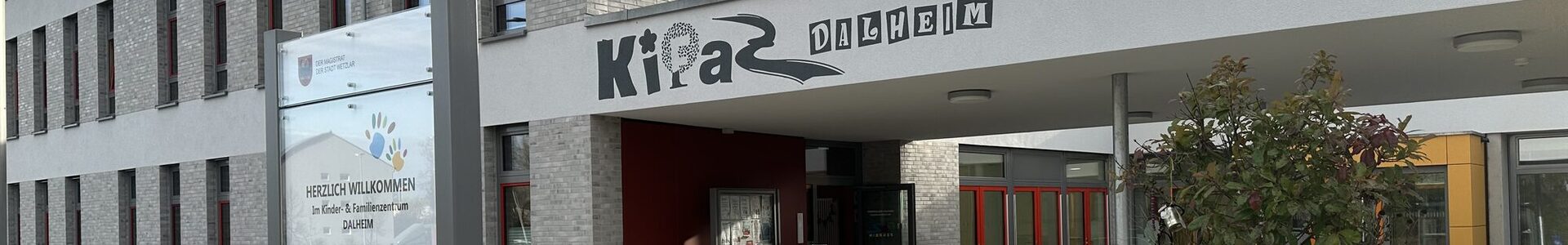 Das Gebäude des Kinder- und Familienzentrums in Dalheim von außen. Im Vordergrund steht ein Schild, dass Besucher des Kinder- und Familienzentrums willkommen heißt. Das Gebäude ist modern und die Außenfassade ist weiß und grau.