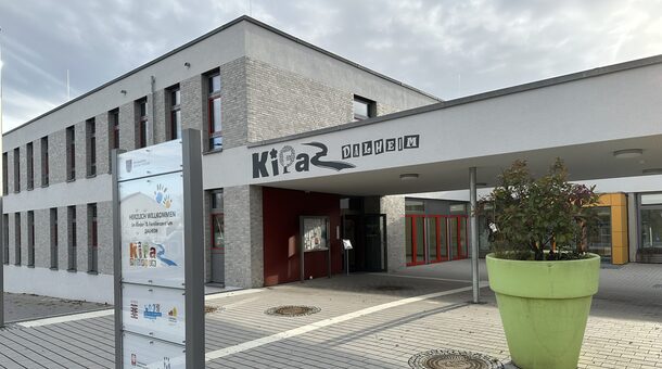 Das Gebäude des Kinder- und Familienzentrums in Dalheim von außen. Im Vordergrund steht ein Schild, dass Besucher des Kinder- und Familienzentrums willkommen heißt. Das Gebäude ist modern und die Außenfassade ist weiß und grau.