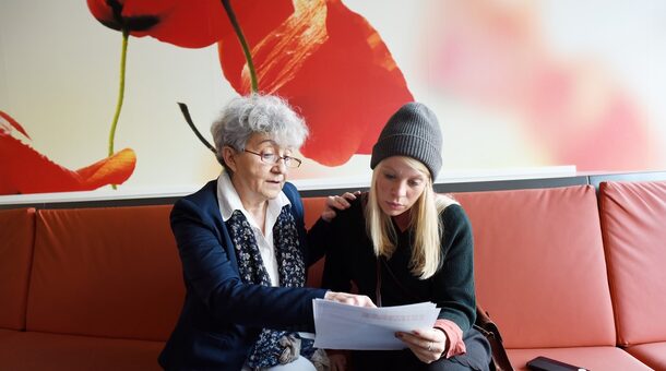 Eine Seniorin und eine Jugendliche auf einem roten Sofa schauen zusammen in einen Text. Sie lesen gemeinsam. Dies nennt man Lautlesetandem.