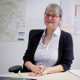 Cornelia Fiebiger, Mitarbeiterin der vhs Leipzig sitzt an ihrem Schreibtisch. An der Wand hinter ihr ist ein Stadtplan zu erkennen.