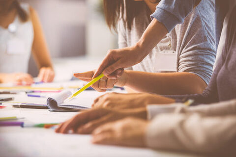 Mehrere Personen sitzen an einem Tisch. Nahaufnahme von Händen. Auf dem Tisch liegen Unterrichtsmaterialien und Stifte.