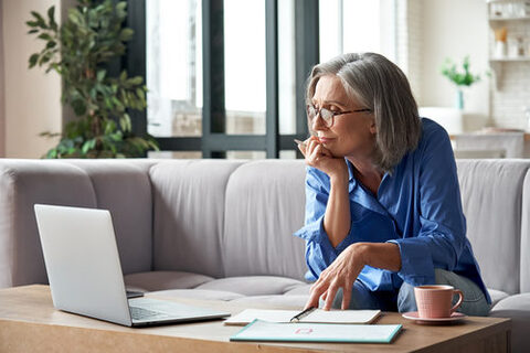 Eine Frau mit Brille und grauen Haaren sitzt auf einem hellen Sofa und beugt sich in Richtung Wohnzimmertisch und schaut dort auf einen Laptop. Auf dem Tisch sieht man außerdem einen Notizblock und eine rosafarbene Kaffeetasse. Im Hintergrund Pflanzen und eine helle Fensterfront.