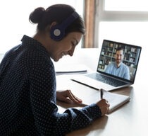 Eine junge Frau mit Kopfhörern sitzt an einem Tisch und schreibt etwas in einen Block, vor ihr ein Laptop auf dem ein Mann vor einem Bücherregal zu sehen ist.