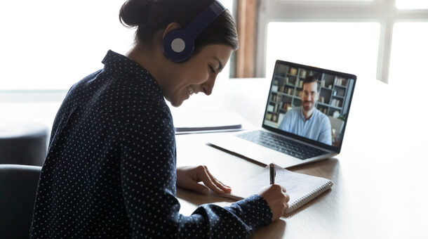 Eine junge Frau mit Kopfhörern sitzt an einem Tisch und schreibt etwas in einen Block, vor ihr ein Laptop auf dem ein Mann vor einem Bücherregal zu sehen ist.