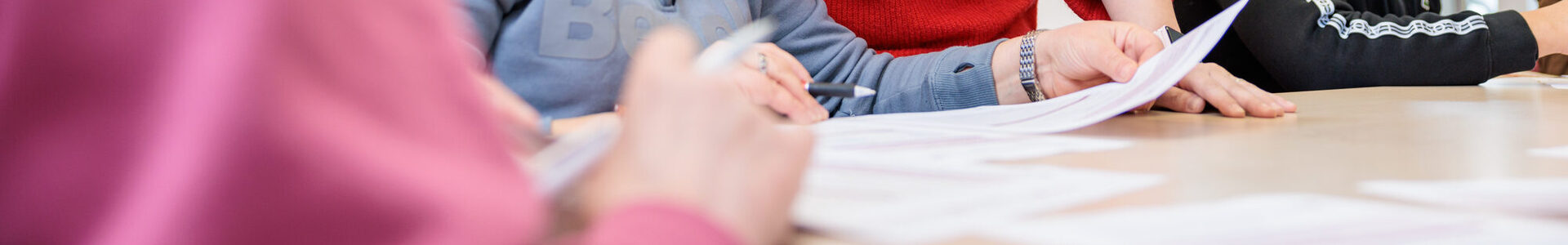 Drei Personen sitzen an einem Tisch und lernen mit Arbeitsblättern. Eine Frau steht uns schaut über die Schulter einer Frau, die am Tisch sitzt.