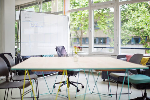 Ein moderner, heller Konferenzraum mit eckigem Tisch, fünf Stühlen und Whiteboard. Durch das Fenster sind Laubbäume und ein weiteres Gebäude zu sehen.