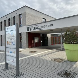 Das Gebäude des Kinder- und Familienzentrums in Dalheim von außen. Im Vordergrund steht ein Schild, dass Besucher des Kinder- und Familienzentrums willkommen heißt. Das Gebäude ist modern und die Außenfassade ist weiß und grau.