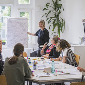Frauen sitzen an einem Tisch und lernen gemeinsam mit zwei Kursleitungen, die an einem Flipchart stehen.