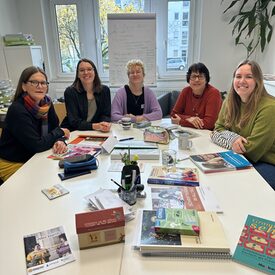 Fünf Frauen sitzen an einem Tisch, auf dem Lernmaterialien liegen, und lächeln in die Kamera.