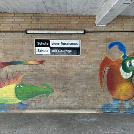 Buntes Graffiti mit Fantasiefiguren schmückt eine Wand an einem Schulhof.